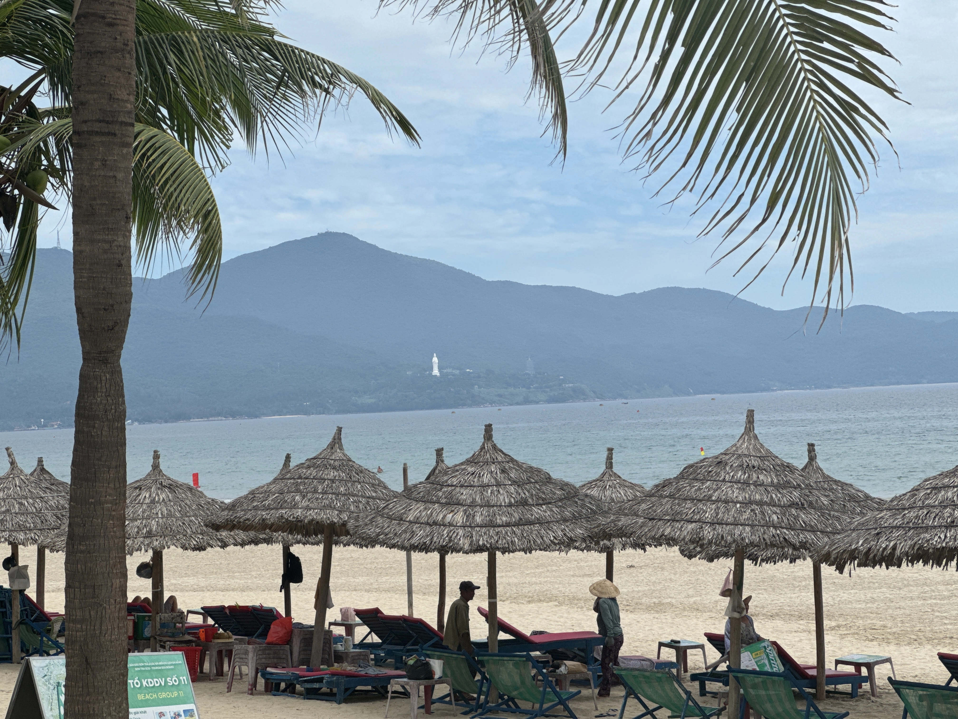 an image of lady buddha in the distance amongst the jungle with bamboo umbrellas providing cover on the beach