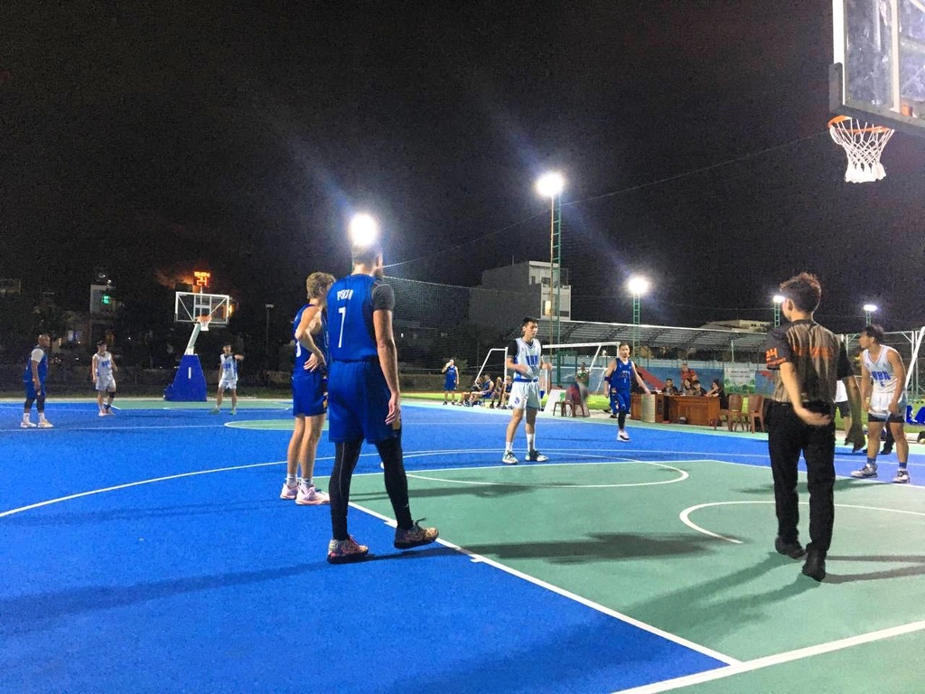 one of my adventures in Vietnam where I played in a basketball tournament with locals, night time lighting, blue jerseys, referee, wide picture of several basketball players from opposing teams